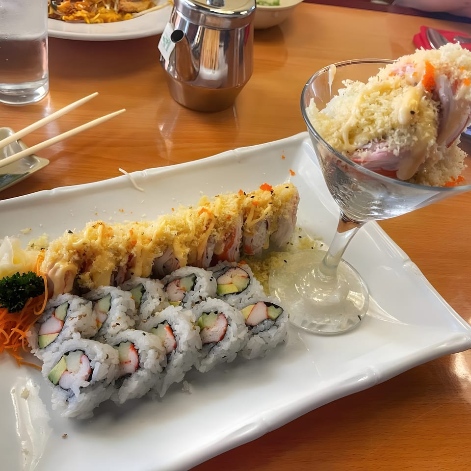 A white platter of sushi rolls, one topped with crispy fried onions, and a martini glass of seafood.