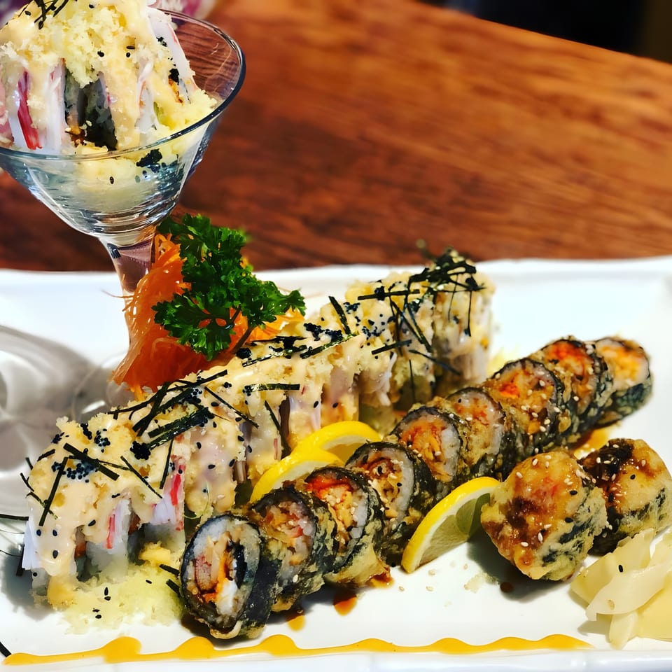 A white plate with various sushi rolls, including deep-fried and sauce-drizzled options, garnished with lemon and ginger.