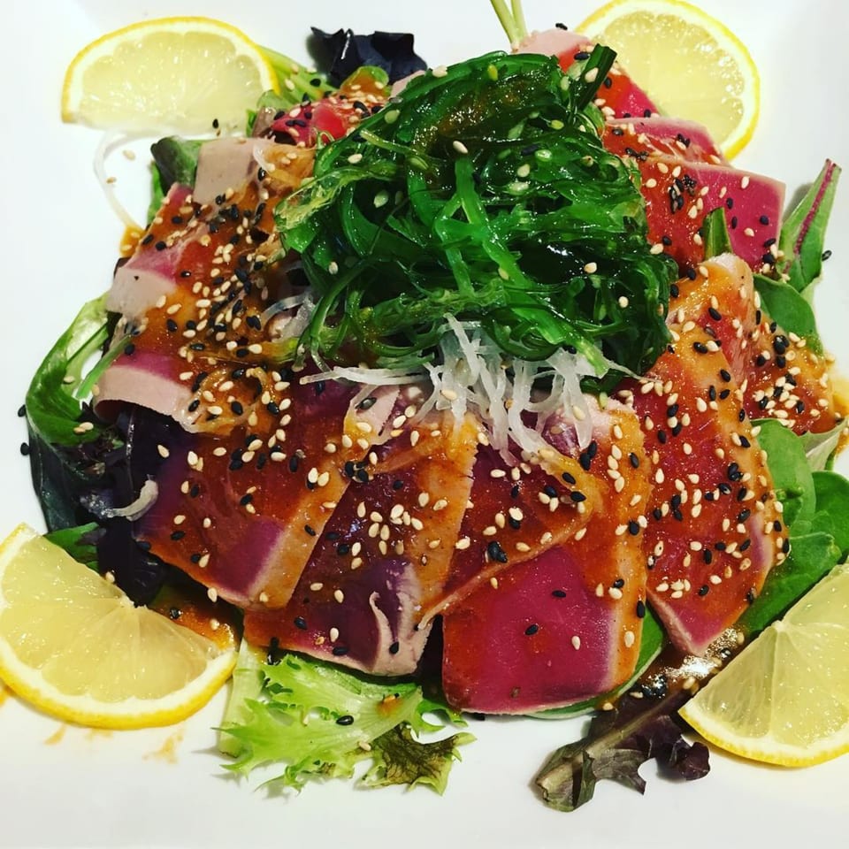 Seared tuna slices with sesame seeds, seaweed salad, and lemon wedges on a bed of greens.